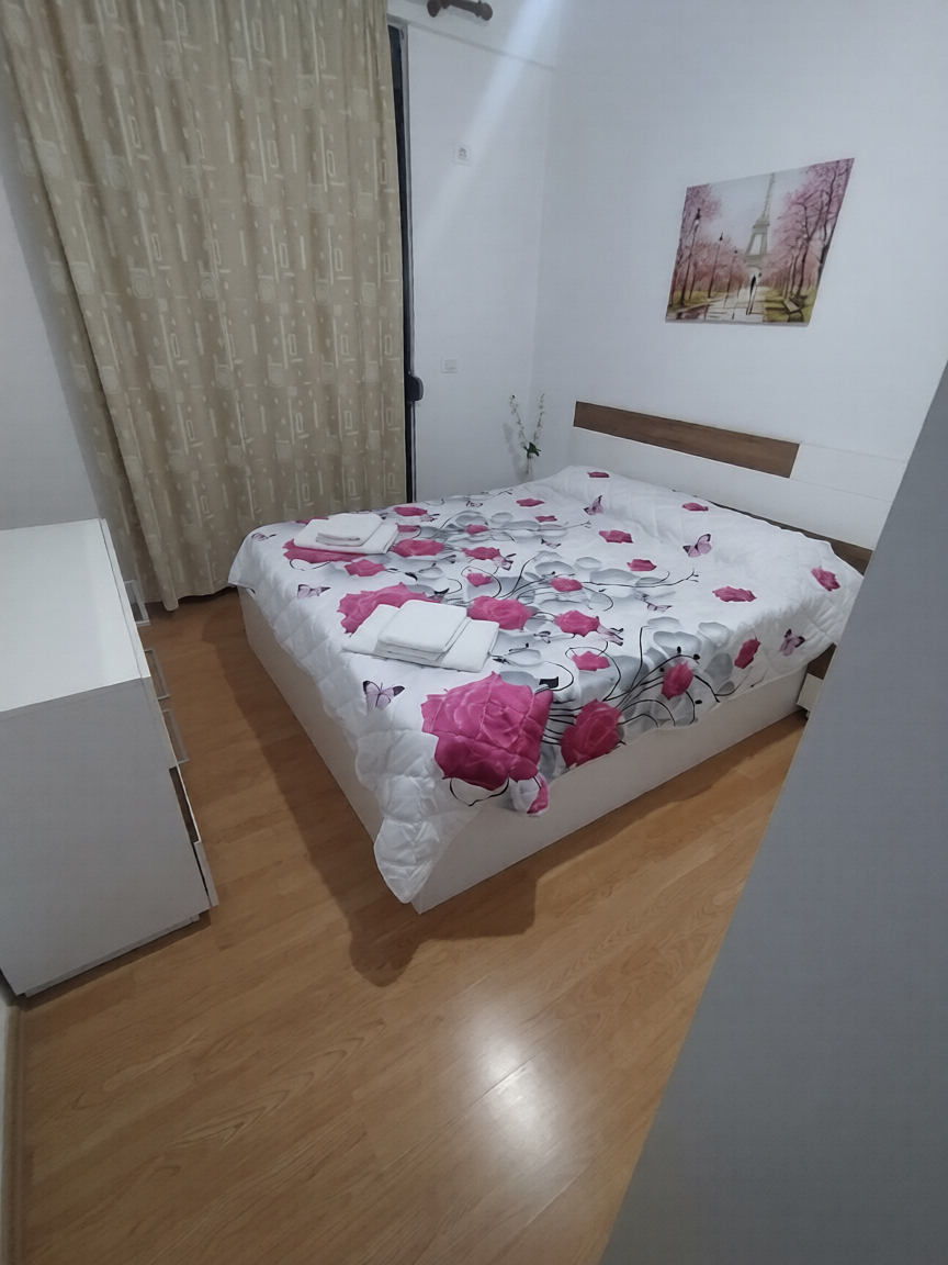 Cozy bedroom with double bed featuring white bedding with pink floral pattern, wooden headboard, beige curtains, wooden floor, wall art, and minimalist decor